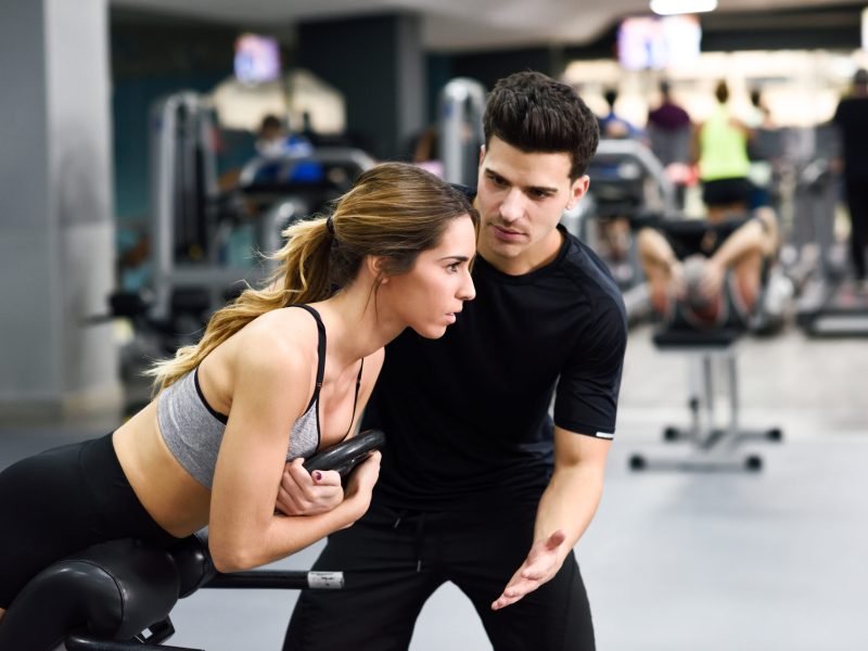 Personal trainer helping young woman lift weights while working out in a gym