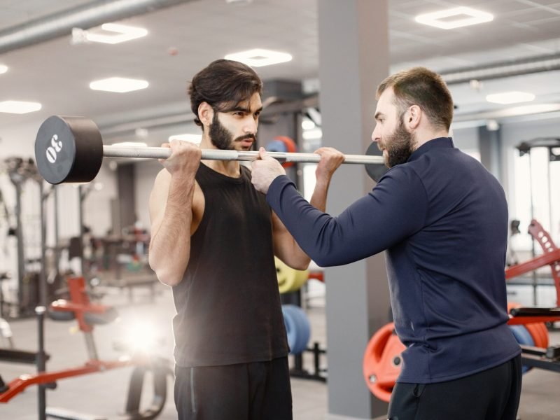 Indian bearded man working out in a modern gym with personal trainer. Man doing excercises on special equipment. Fit man wearing black t-shirt.